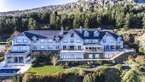 Una casa grande de varios pisos con un balcón destacado, rodeada de abundante vegetación y un paisaje montañoso.