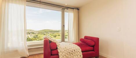 Ein Zimmer mit einem roten Sofa, einem großen Fenster mit Vorhängen und Blick auf eine grüne Landschaft.