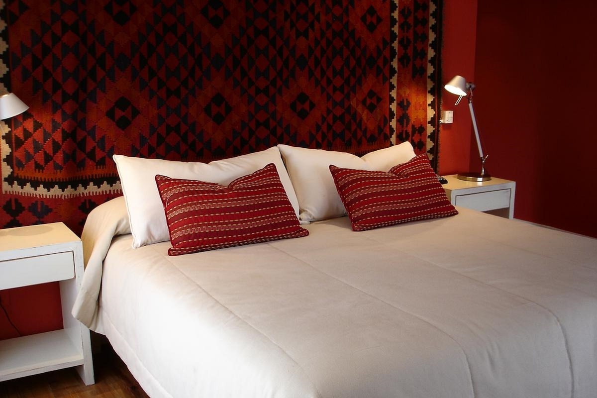 A neatly made bed with white bedding and two red patterned pillows, a bedside lamp, and a patterned wall hanging.