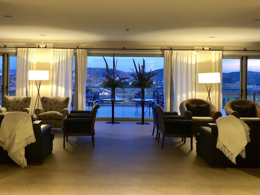 A spacious living room with a large window offering a view of a mountainous landscape. There are two round tables with chairs, a sofa, and two floor lamps.