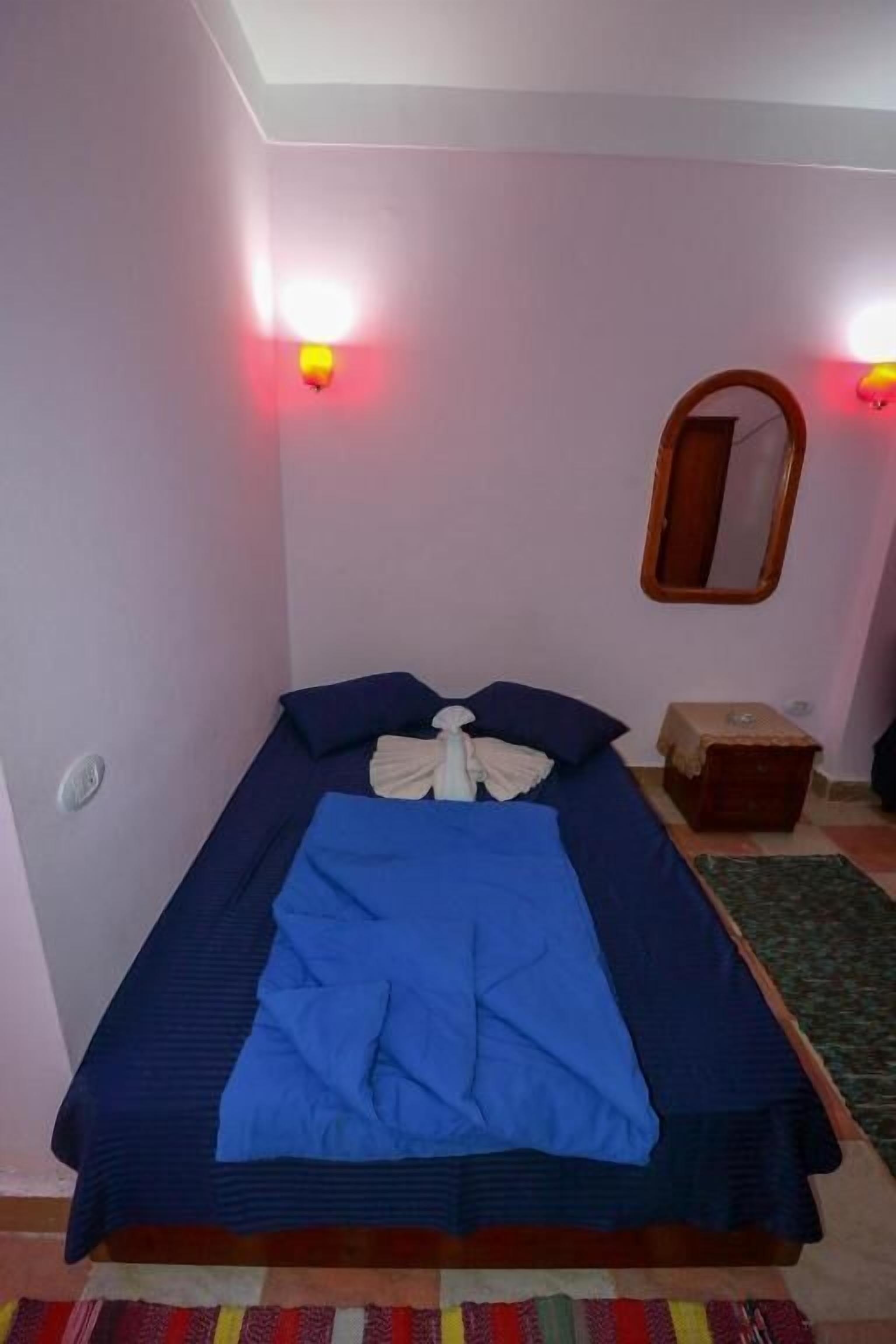 A single bed with a blue blanket and pillows, a small bedside table, a mirror on the wall, and a red wall light.