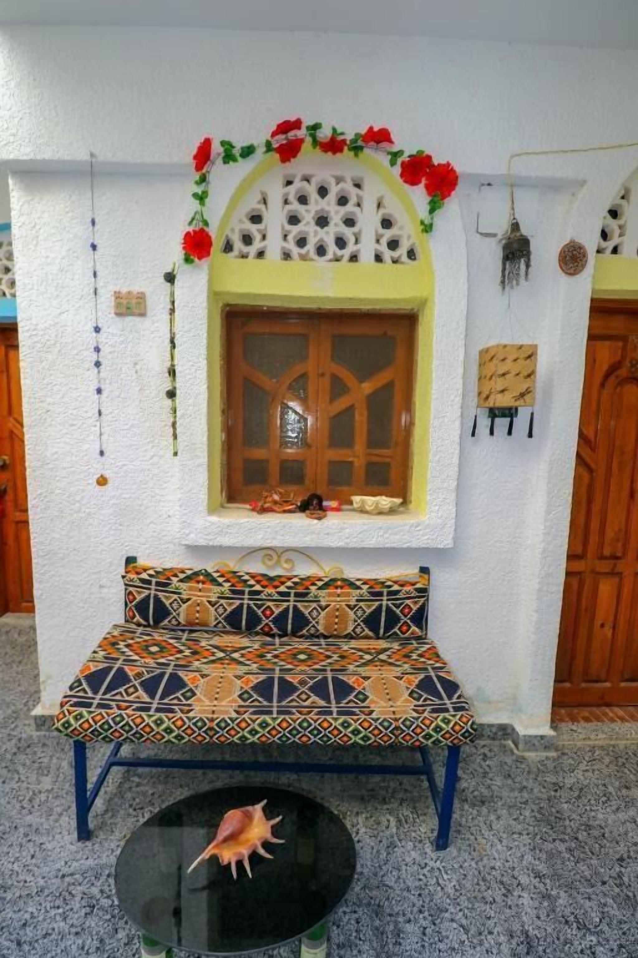 A room with a patterned bench, a small table with a shell, a window with a wooden frame, and a decorative archway.