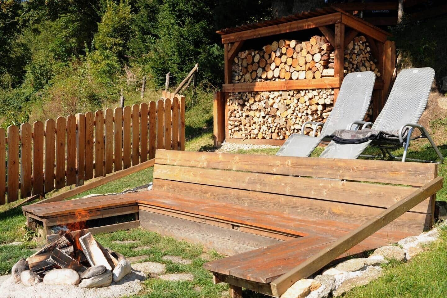 Una terraza de madera con un brasero, sillas de descanso y una pila de leña.