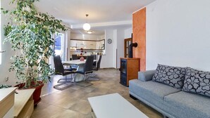 A modern living room with a grey sofa, a dining table with chairs, a large potted plant, and a kitchen in the background. - Holiday apartments Carmen / Carmen Garden (Pula)