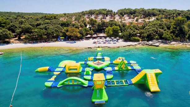 An aerial view of a beach with inflatable water play structures in the water and a coastal town in the background. - Holiday apartments Carmen / Carmen Garden (Pula)