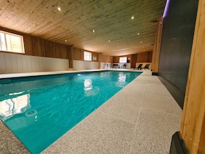 An indoor swimming pool with a wooden ceiling and tiled floor, surrounded by a lounge area with chairs and a counter. - Fantastic panoramic views, beautiful Peak District Cottage with Indoor Pool (High Peak)