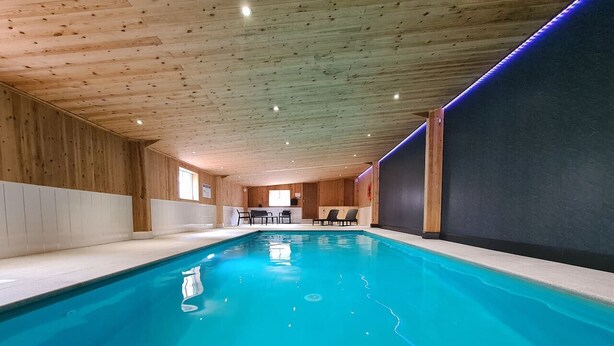 Una piscina interior con paredes y techo de madera, agua azul cristalina y un área de estar con sillas y mesas.