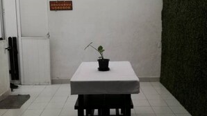 Uma pequena mesa quadrada com toalha branca e um vaso de planta em cima, colocada num quarto com piso de azulejos, uma porta branca e uma parede verde ao fundo.