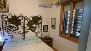 Ein Schlafzimmer mit einem Bett, einem dekorativen Metall-Kopfstück, einem Nachttisch mit Lampe, einem Fenster mit Jalousien und einem gerahmten Bild an der Wand.