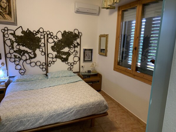 A bedroom with a bed, a decorative metal headboard, a bedside table with a lamp, a window with blinds, and a framed picture on the wall. - Double room with bathroom 150 meters from the beach (Quartu Sant'Elena)