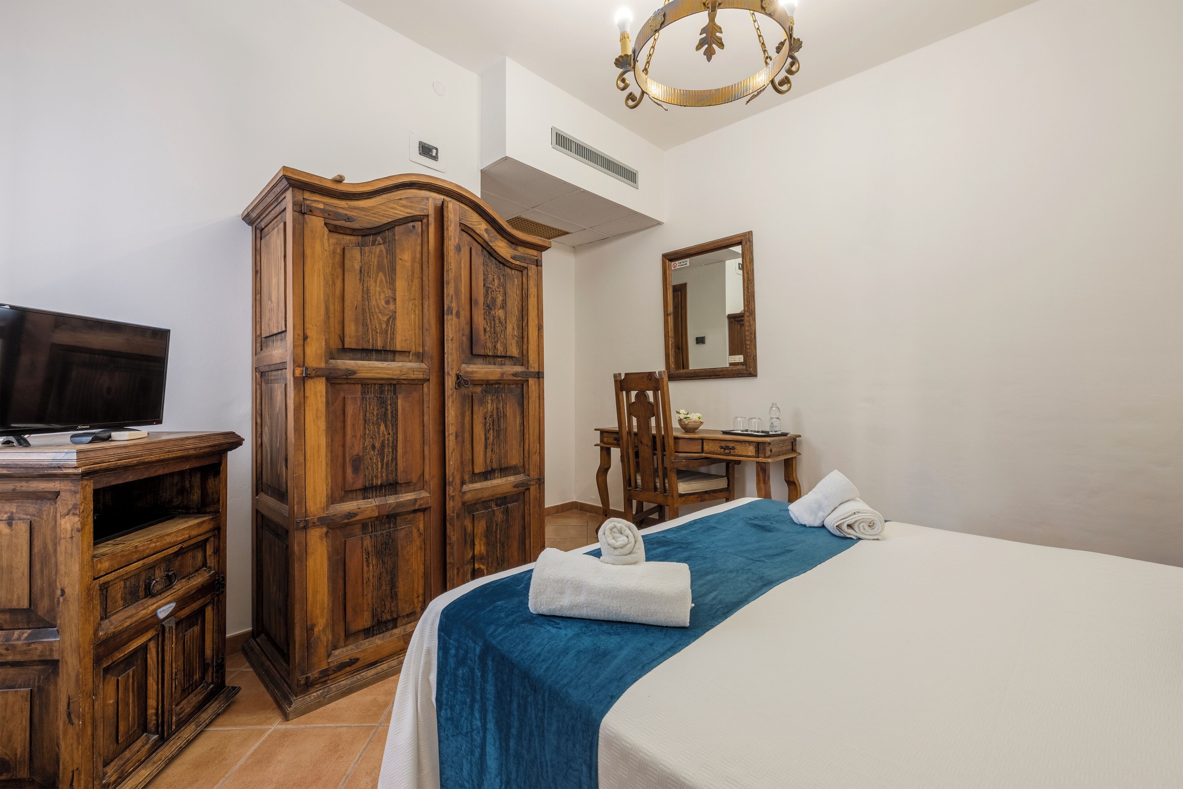 A hotel room with a wooden wardrobe, a bed with a blue bedspread, a television on a wooden cabinet, a desk with a chair, and a mirror on the wall.