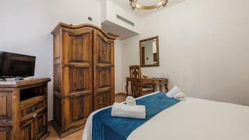 Ein Hotelzimmer mit Holzschrank, einem Bett mit blauer Bettdecke, einem Fernseher auf einer Holzkommode, einem Schreibtisch mit Stuhl und einem Spiegel an der Wand.