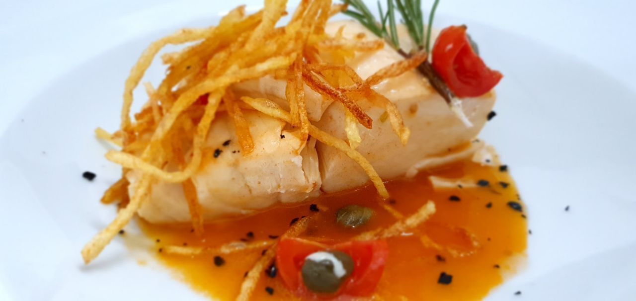 A dish of fish with crispy skin, garnished with herbs and a red flower, served on a white plate.