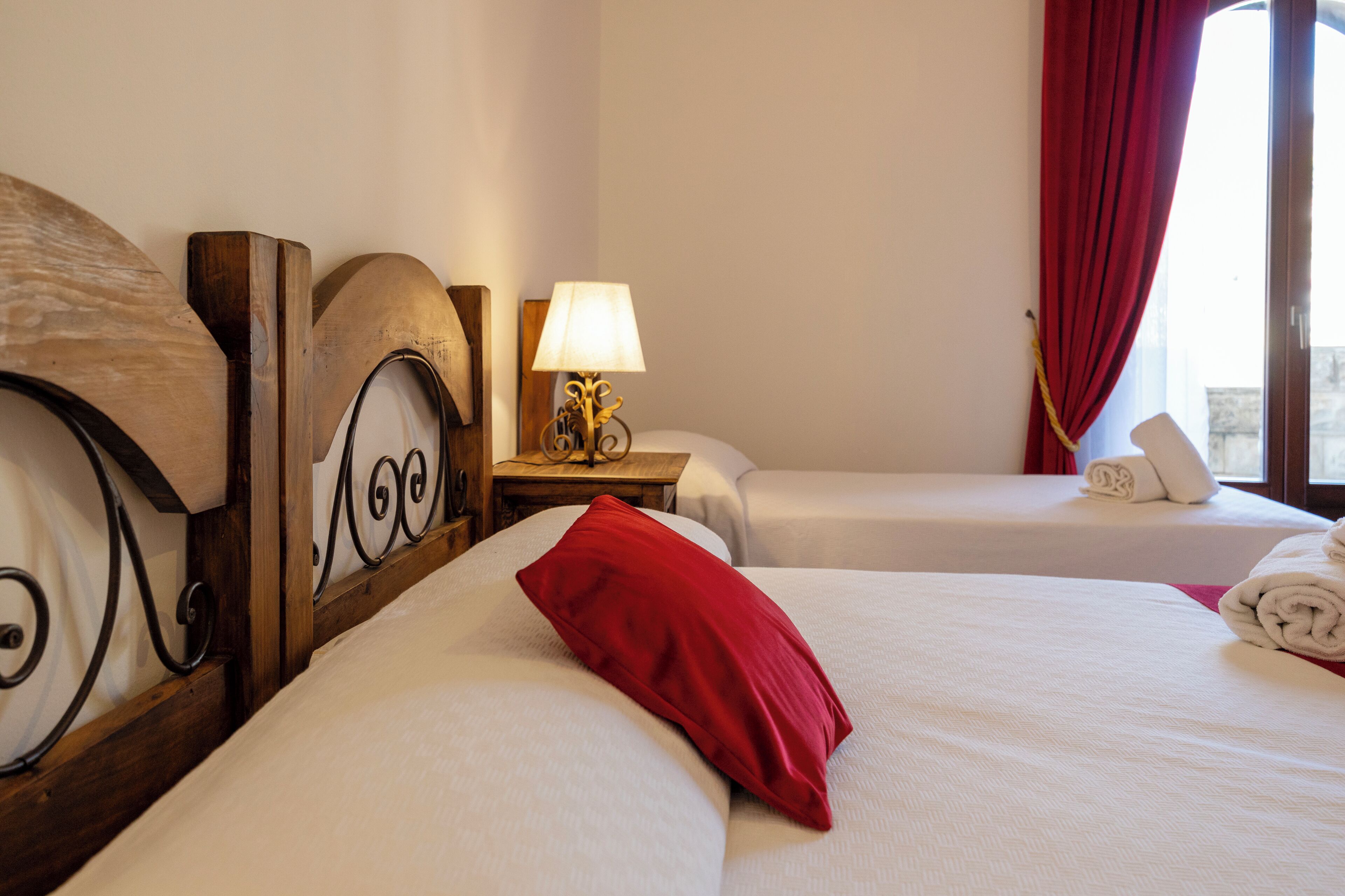 Habitación de hotel con dos camas, cabecera de madera, mesita de noche con lámpara y ventana con cortinas rojas.