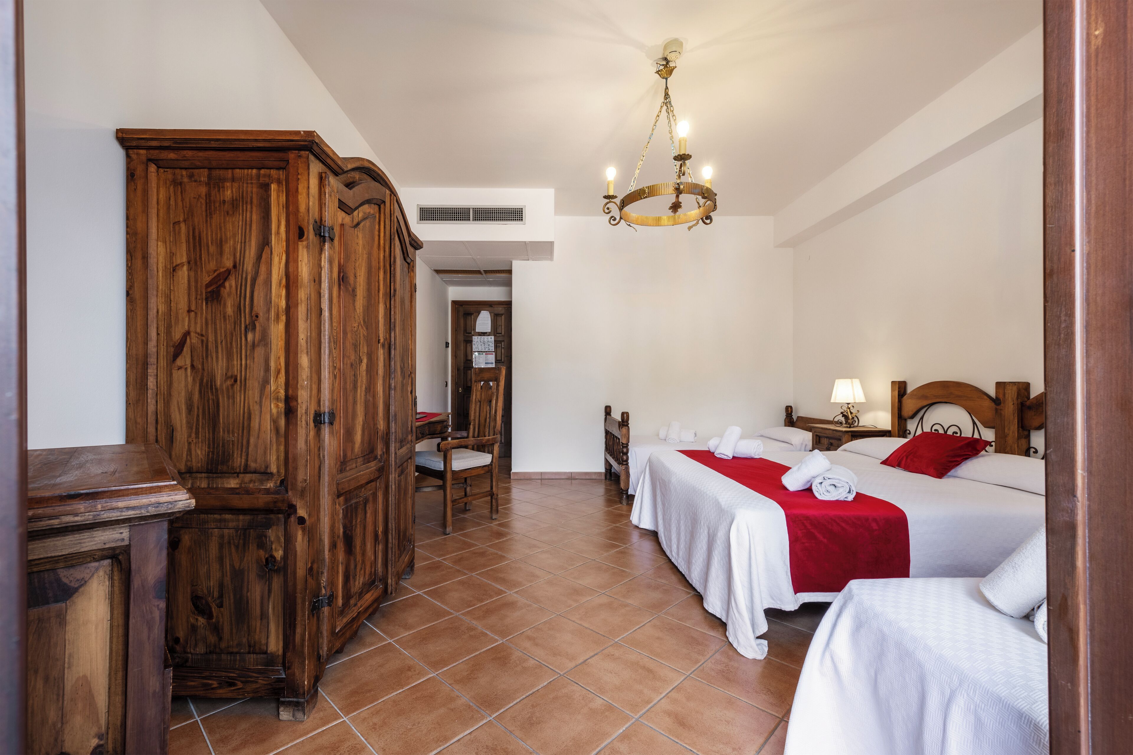A hotel room with two beds, a wooden wardrobe, a chandelier, and a tiled floor.