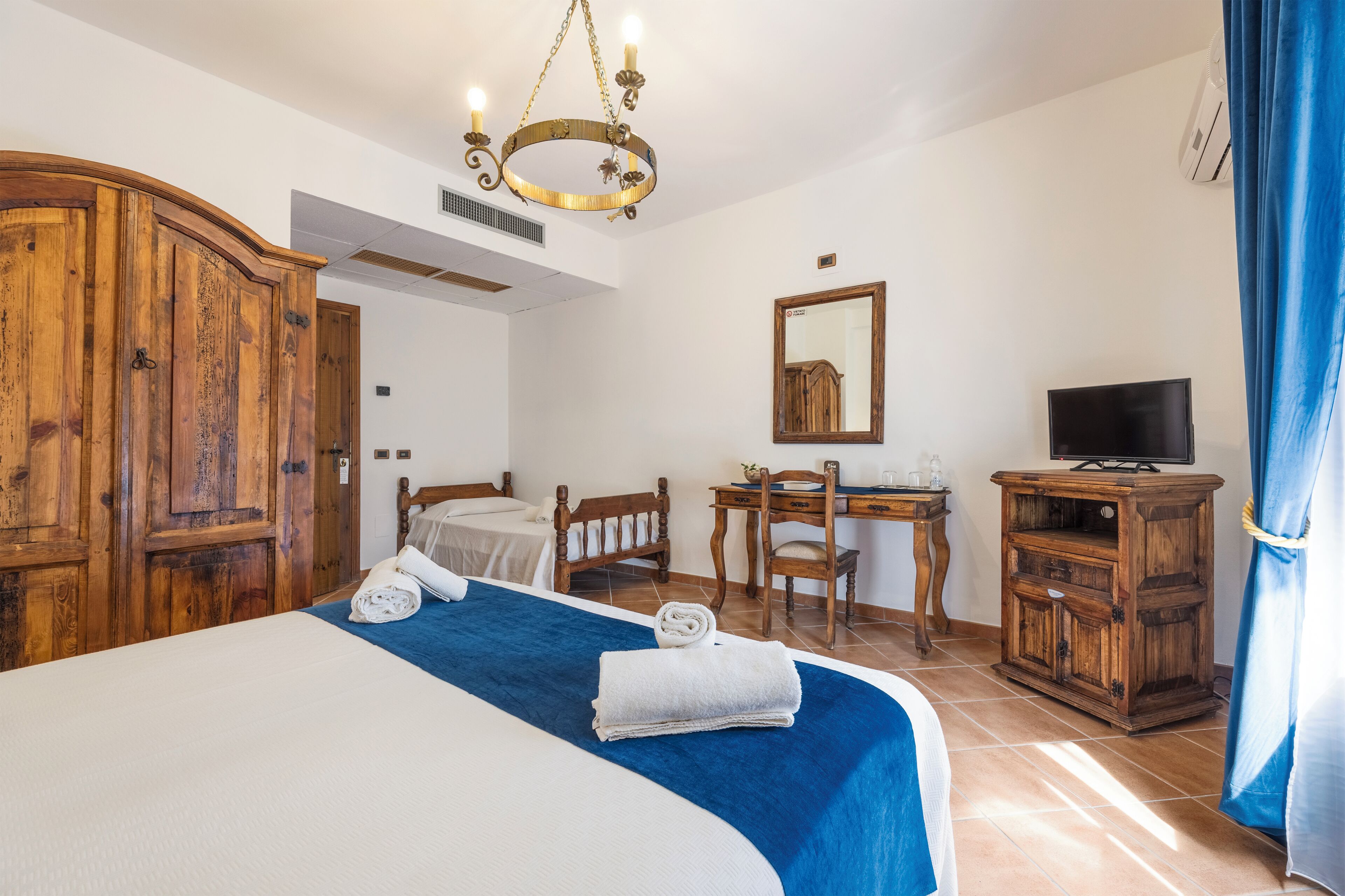 A hotel room with a large bed, a wooden wardrobe, a desk with a chair, a television, and a mirror on the wall.