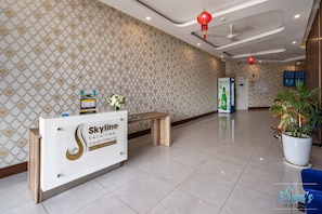 Un área de recepción con un mostrador identificado como 'Skyline Services Apartments' y un número de contacto. Se observa un banco, una planta en maceta y una pantalla de televisión instalada en la pared.