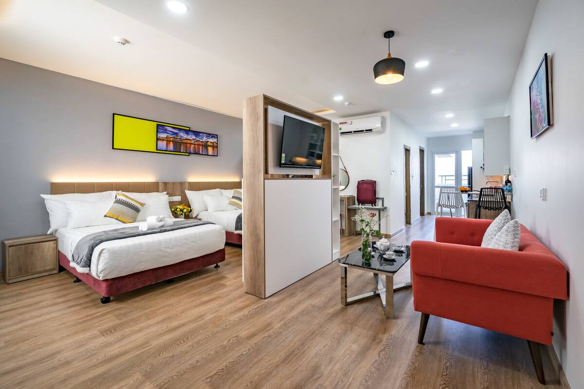 A modern hotel room with a large bed, a red armchair, a TV, and a dining area in the background.