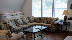 Un salon comprenant un canapé d’angle, une table basse en verre, une petite table d’appoint et une lampe. De grandes fenêtres sont équipées de stores, et on y trouve également un meuble blanc et une cheminée.