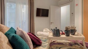 Una habitación de hotel con una cama, un televisor, una mesa con hervidor y tazas, y una puerta con un letrero rojo en forma de 'X'.