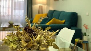 Una sala de estar con un sofá color turquesa, cojines amarillos, una mesa de centro de vidrio con flores secas y una lámpara.