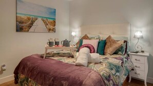 Un dormitorio con cama, mesitas de noche, lámpara, un cuadro y una mesa con objetos encima.