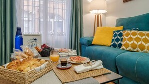 Una sala de estar acogedora con un sofá color turquesa, una lámpara y una mesa para el desayuno con croissants, jugo de naranja y una taza de café.