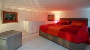 Une chambre avec un grand lit, une table de chevet, un petit banc et deux tableaux encadrés au mur.