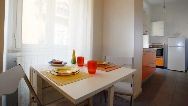 Un coin repas avec une table dressée pour deux personnes, une cuisine en arrière-plan et une fenêtre ornée de rideaux légers.