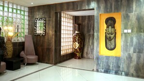 A lobby with a dark tiled wall, a large vase with floral patterns, a chair, and a decorative mirror. - La Cour Hotel Apartments (Lagos)