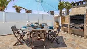 Een terras buiten met een ronde tafel, zes houten stoelen, een blauwe parasol en een stenen barbecue.