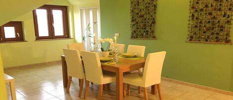 Un coin repas avec une table en bois dressée pour quatre personnes, entourée de chaises. On y trouve une fenêtre avec des stores, un mur vert agrémenté d’un panneau décoratif et un sol carrelé.