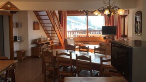 Un salon chaleureux avec un escalier en bois, un coin repas avec des meubles en bois, un téléviseur et une vue sur un paysage montagneux.