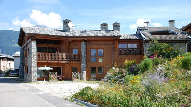 Un chalet en bois doté d’un balcon, de cheminées en pierre et d’un jardin où poussent différentes variétés de fleurs.