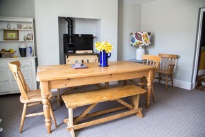 A wooden dining table with benches, a blue vase with yellow flowers, and a black stove in the background. - Delightful, Dog-Friendly Cottage in Millbrook, Cornwall near the coast (Millbrook)