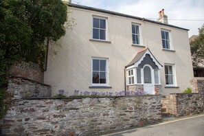 A two-story building with a stone wall and a small garden with purple flowers. - Delightful, Dog-Friendly Cottage in Millbrook, Cornwall near the coast (Millbrook)