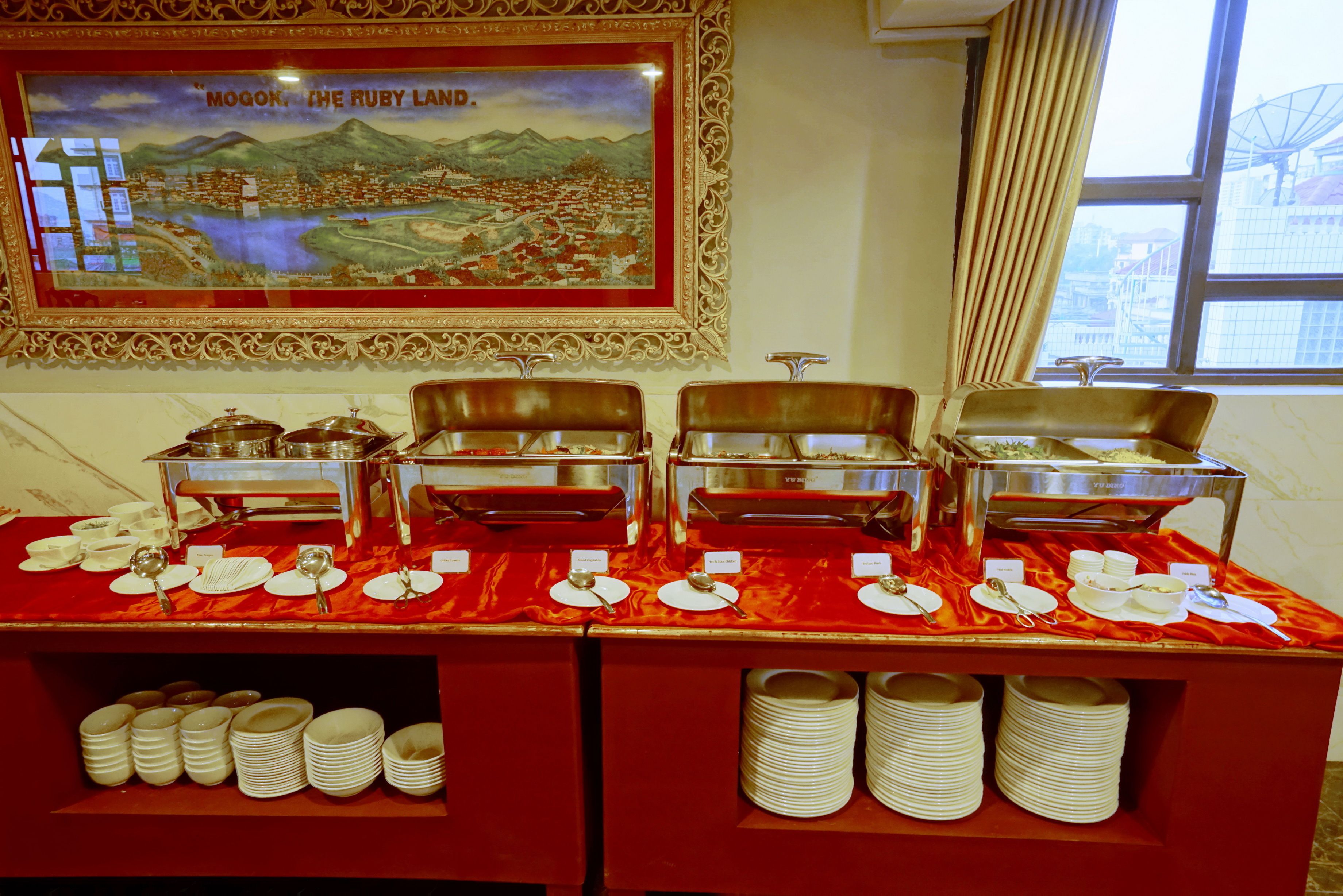 A buffet setup with multiple serving stations, a framed cityscape painting, and a window with a view of buildings.
