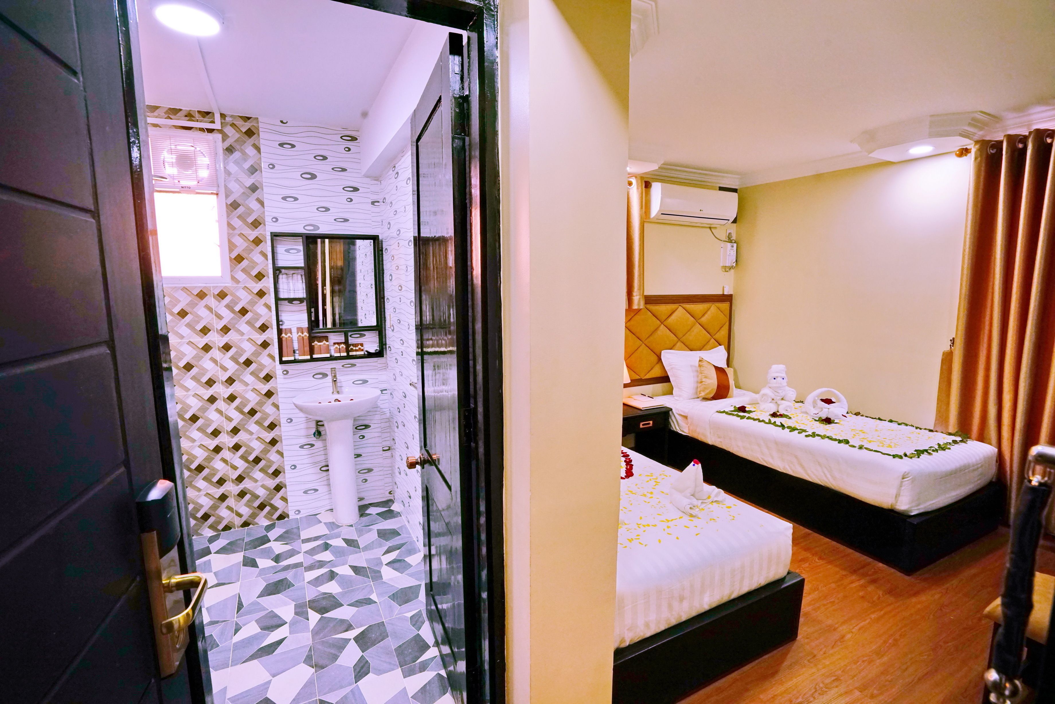 A hotel room with a tiled floor, a bed with a white bedspread, a bedside table, and a wall-mounted air conditioning unit.