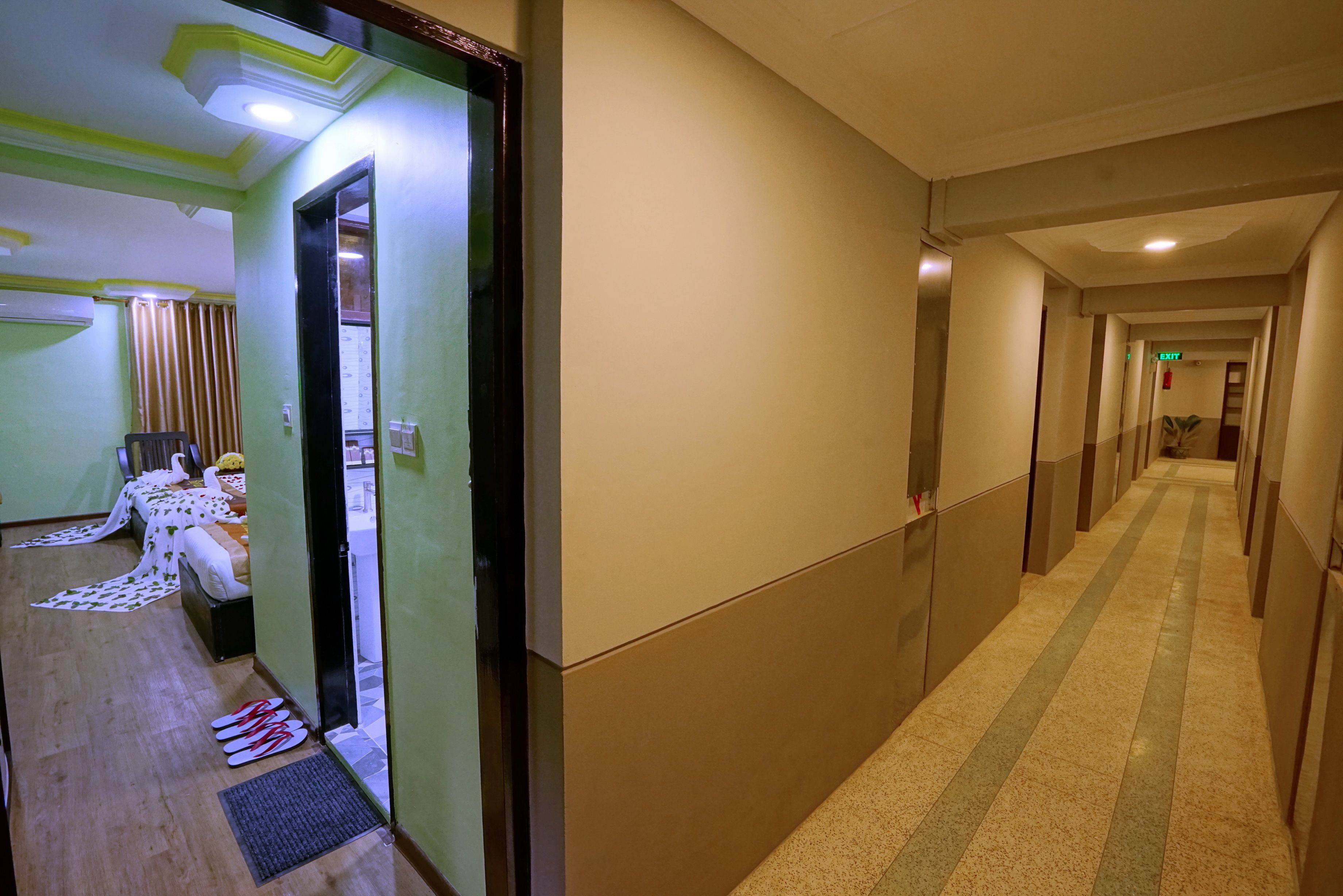 A hotel hallway with a carpeted floor, a door leading to a room, and a bench with a towel and slippers.