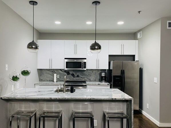 A modern kitchen with white cabinets, a stainless steel refrigerator, a microwave, and a marble island with bar stools.
