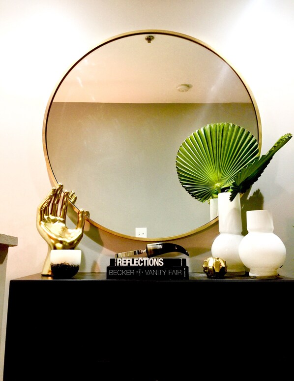 Une coiffeuse avec un miroir rond, une sculpture dorée représentant une main, un éventail et un vase avec une plante.