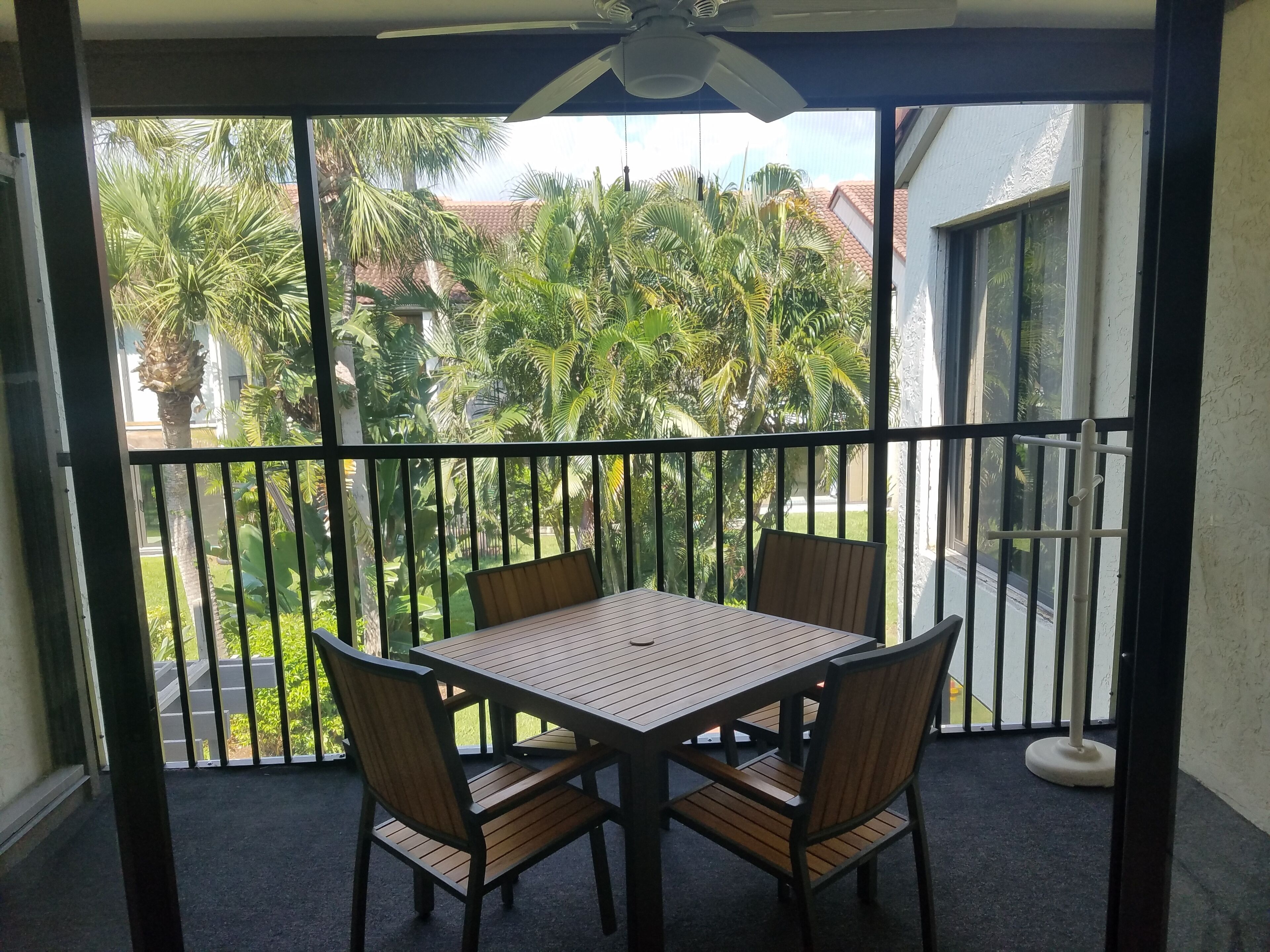 Un balcon avec une table et des chaises, un ventilateur de plafond et une vue sur un jardin planté de palmiers.