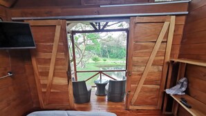 Quarto de madeira com uma cama, uma TV fixada na parede e vista para um jardim através de portas abertas.