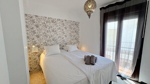 A bedroom with a floral wallpaper, a bed with white linens, a nightstand with a lamp, and a balcony with a view. - AP290 - Casa Olivia (Nerja)