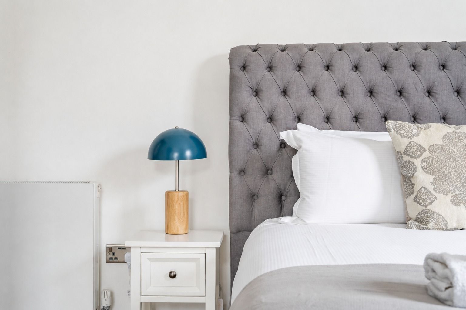 A neatly made bed with a tufted headboard, a bedside lamp, and a small bedside table with a drawer.