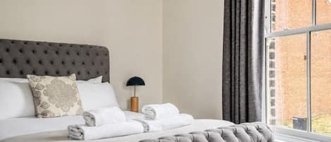 A neatly made bed with a tufted headboard, a bedside lamp, and a view of a brick building through a window.