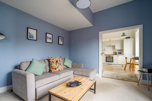 Un salon avec un canapé gris, une table basse en bois et une cuisine visible à travers une porte ouverte.
