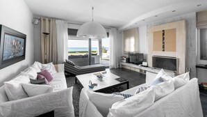 A modern living room with a white sofa set, a coffee table, and a TV. There is a large window with a view of the ocean.