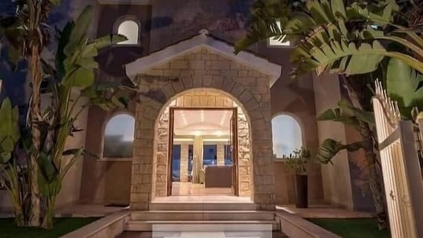 Un bâtiment en pierre avec une entrée centrale, flanqué de grandes fenêtres et entouré de verdure.