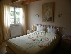 A bedroom with a bed, a window with curtains, a radiator, a nightstand, and a painting on the wall.